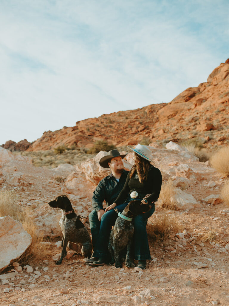 Wedding Photographers Reno capture couple sitting together on rocks embracing with dogs running around at Red Rocks