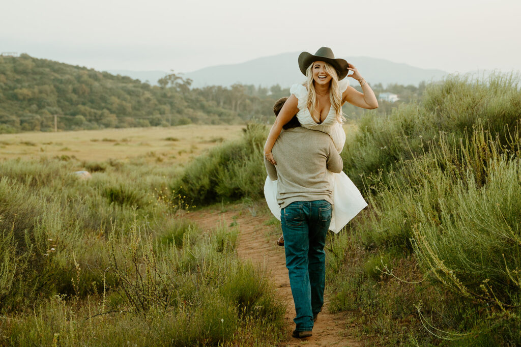 Reno wedding photographer captures man lifting woman on back while she wears cowboy hat during summer engagement photos